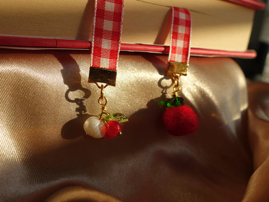 Bookmark with red and white checkered ribbon, gold accents, beaded cherries, beaded tomato, and a pearl on a textured surface